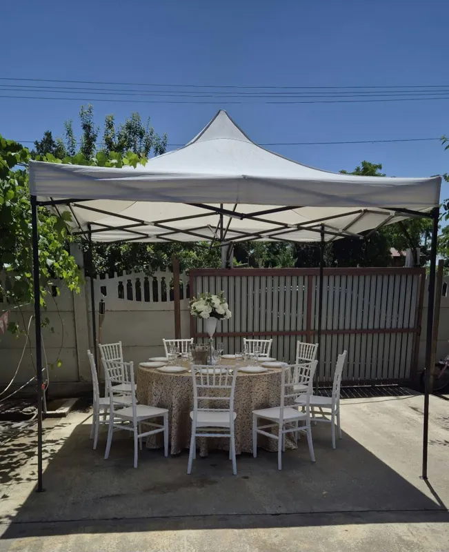 Pavilion alb cu masă rotundă acoperită cu față de masă aurie, flori și scaune albe